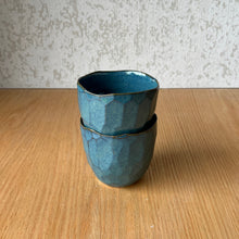 Load image into Gallery viewer, Two stacked ceramic cups with a textured, faceted geometric design in a teal blue glaze, set on a light wood tabletop against a textured gray wall.